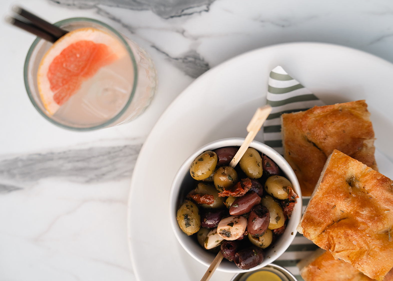Bowl of olives with bread and a glass of pink grapefruit drink on a marble surface